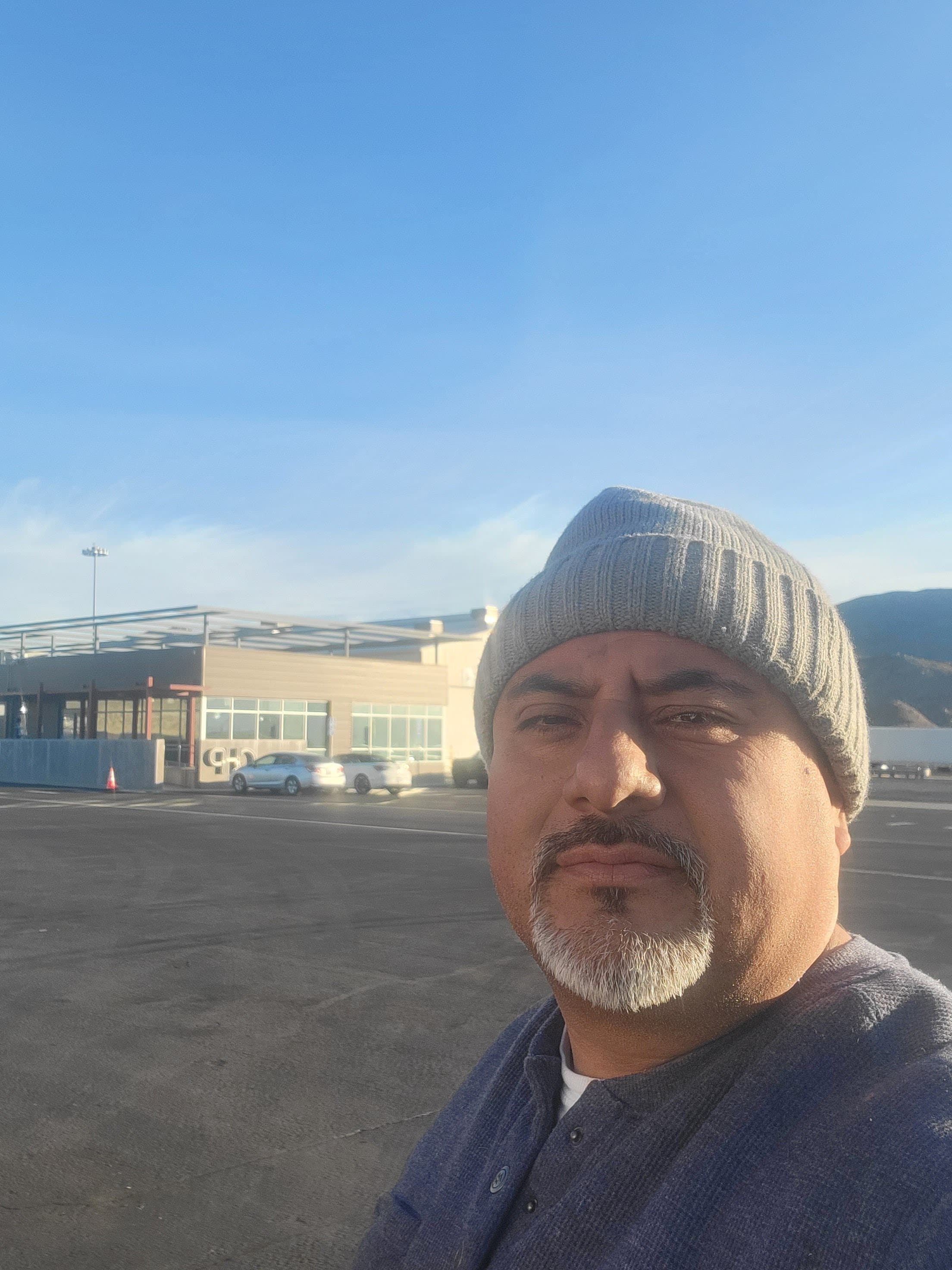 Man wearing a grey beanie and goatee posing outdoors in a sunny parking lot.