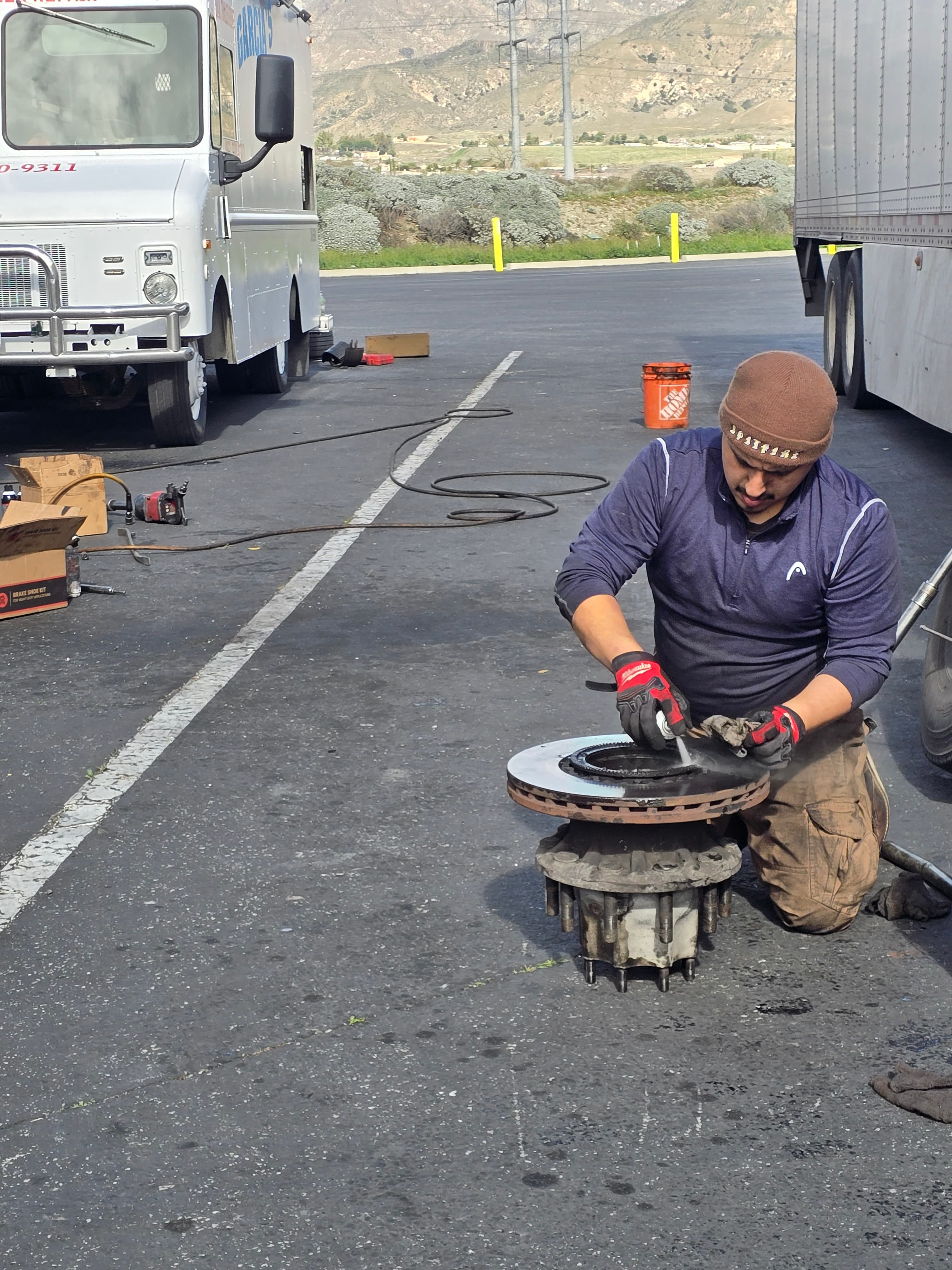 A mechanic in a beanie and gloves sprays a large brake rotor on the asphalt.